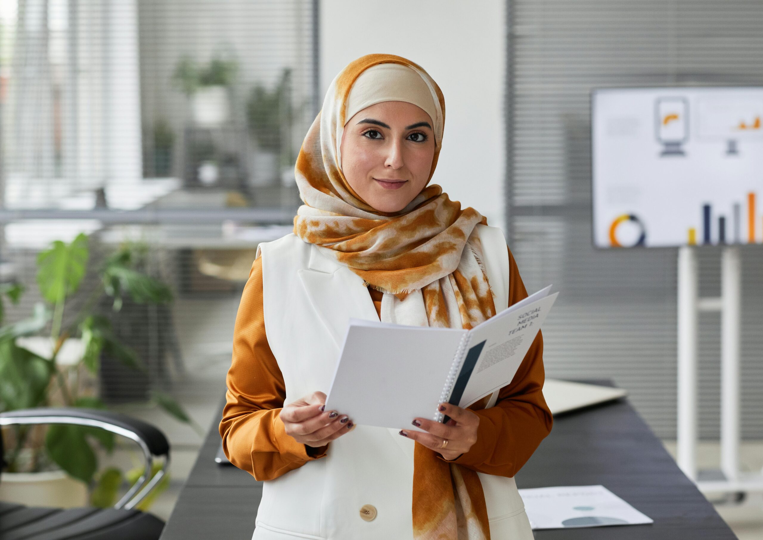 A professional Muslim woman in an office setting, confidently holding documents and smiling.
